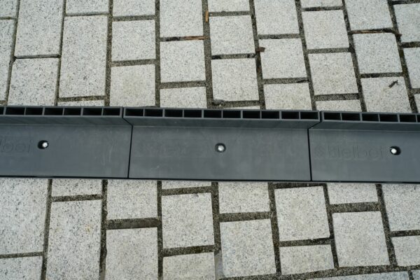 A linear drainage channel with a black grate cover running through a pavement made of square stone tiles.