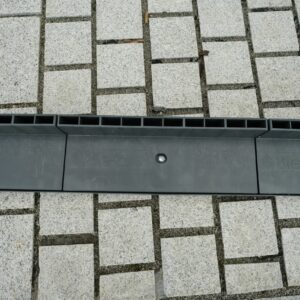 A linear drainage channel with a black grate cover running through a pavement made of square stone tiles.