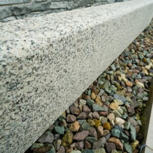 A diagonal angle view of a polished granite curb next to a bed of multicolored pebbles with a stone wall in the background.