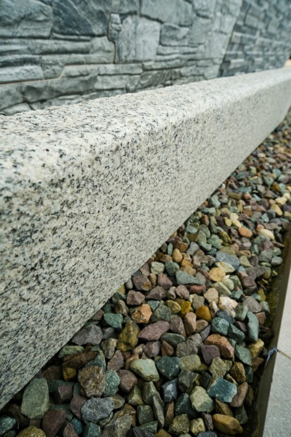 A diagonal angle view of a polished granite curb next to a bed of multicolored pebbles with a stone wall in the background.