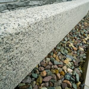 A diagonal angle view of a polished granite curb next to a bed of multicolored pebbles with a stone wall in the background.