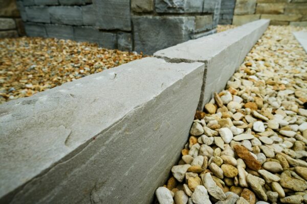 A close-up view of a modern landscaping design featuring linear concrete bench edges with a pebble ground cover, set against a grey stone wall in the background.