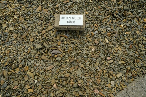 A concrete block with the label "BRONZE MULCH 40MM" sits on a bed of small, multicolored stones and fragments.