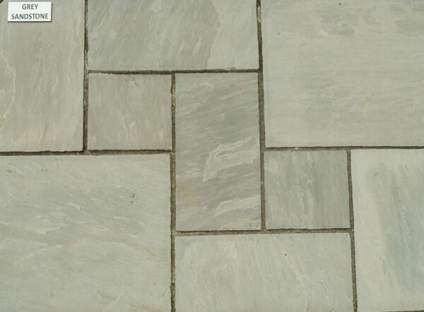 A variety of grey sandstone tiles laid out on the ground, showcasing different sizes and shapes, with a labeled sign "GREY SANDSTONE" placed on one of the tiles.