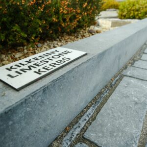 A sign reading "Kilkenny Limestone Kerbs" placed on a stone curb next to a paved pathway and a shrubbery bed.