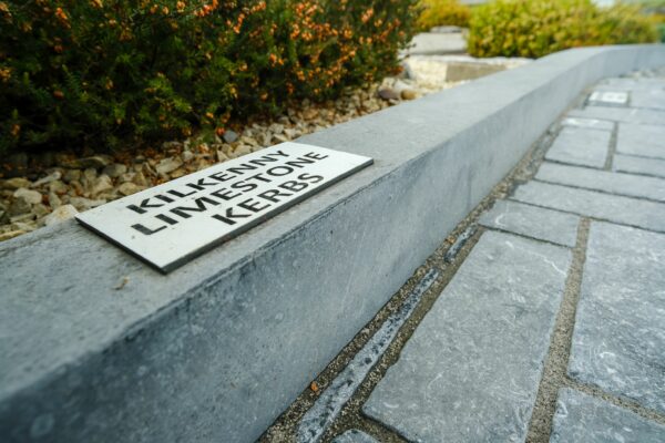 A sign reading "Kilkenny Limestone Kerbs" placed on a stone curb next to a paved pathway and a shrubbery bed.