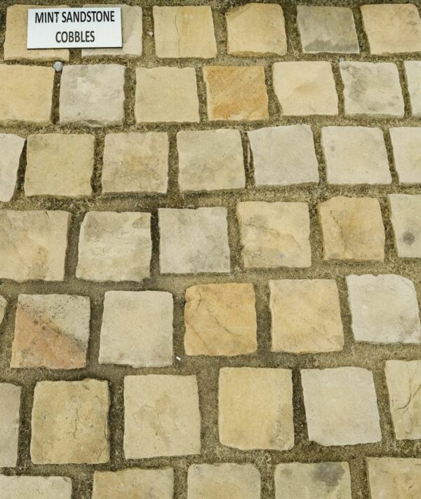 A stone pavement displaying mint sandstone cobbles, arranged in an interlocking pattern with a strip of grey stones, accompanied by a label indicating their type and bordered by pebbles and a concrete edge.