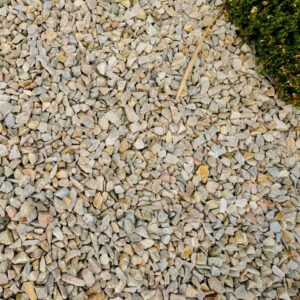 A large number of pink quartz stones labeled "PINK QUARTZ 20MM" spread out on the ground next to a concrete curb and a small green bush.