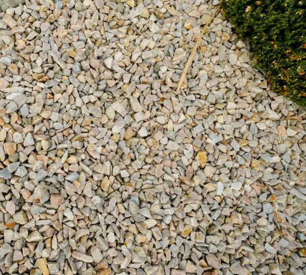 A large number of pink quartz stones labeled "PINK QUARTZ 20MM" spread out on the ground next to a concrete curb and a small green bush.