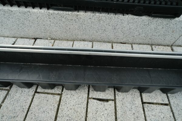 A ground-level view of a drainage system next to a pavement, featuring a metal grate with parallel bars over a black trough, adjacent to a textured stone curb.