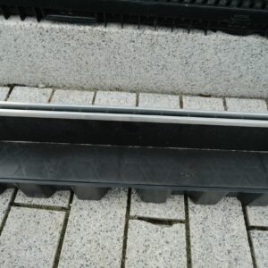 A ground-level view of a drainage system next to a pavement, featuring a metal grate with parallel bars over a black trough, adjacent to a textured stone curb.