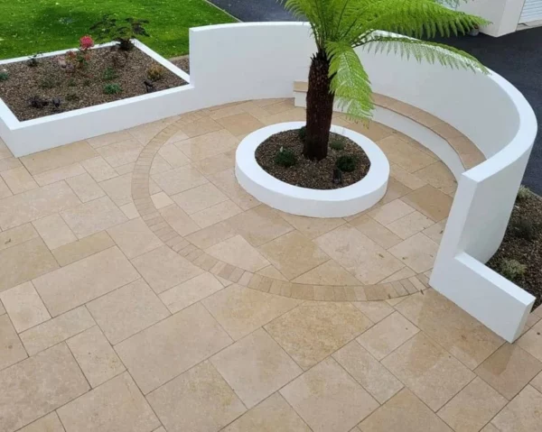 A modern outdoor patio with beige tiles, featuring curved white walls surrounding landscaped areas with plants and a central palm tree.