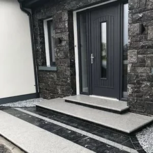 A front entrance of a modern house featuring a dark grey door with panels, silver hardware, and a narrow sidelight. The facade is adorned with dark stone cladding. There are three steps leading to the door with a black and white tiled design on the porch floor.