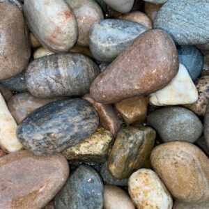 A close-up photo of smooth, multi-colored pebbles, possibly wet, showcasing a variety of earth tones.