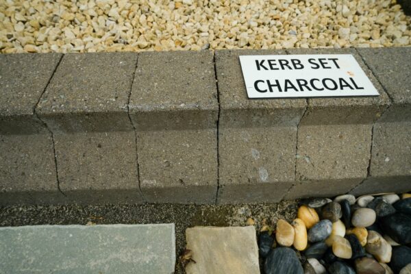 A row of gray kerbstone blocks with a sign reading "KERB SET CHARCOAL" placed on top, surrounded by multi-sized gravel and pebbles on either side.