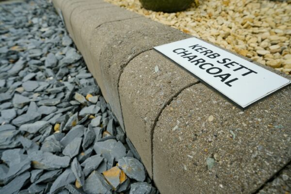 A close-up view of a curved concrete kerb with a sign that reads "KERB SETT CHARCOAL" resting against it, with a background featuring scattered gray slate chippings to the left and beige pebbles to the right.