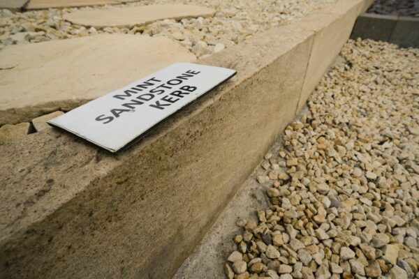 A close-up image of a sandstone kerb with a label that reads "MINT SANDSTONE KERB" placed on it, surrounded by smaller loose stones.