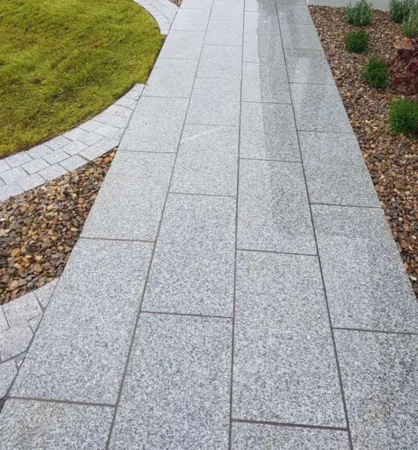 A well-maintained garden pathway made of large rectangular grey paving stones, flanked by a neatly trimmed grass lawn on the left and decorative gravel with small shrubs on the right.