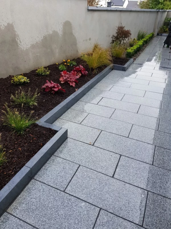 A well-maintained garden pathway with large gray paving stones and raised beds lined with dark metal edging. The beds are planted with a variety of shrubs and colorful flowers against a plain concrete wall.