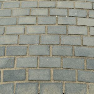 A neatly arranged black limestone sett pavement with a label on top, bordered by gravel and different types of paving stones on either side.