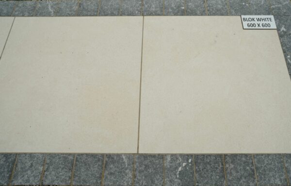 A sample display of two large, square, beige paving slabs labeled "BLOK WHITE 600 X 600," edged with a row of smaller, rectangular grey slabs, all on a bed of small pebbles.