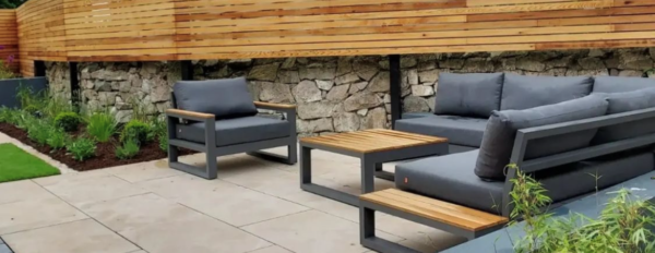 An outdoor seating area with modern gray furniture, including a sofa and armchairs with cushions, and a wooden coffee table, set on a paved patio. The patio is bordered by a curved wooden fence and stone retaining wall with landscaped garden beds.