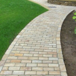 Alt text: A curving pathway made of interlocking bricks leads through a garden area, with grass on one side and bare soil on the other.