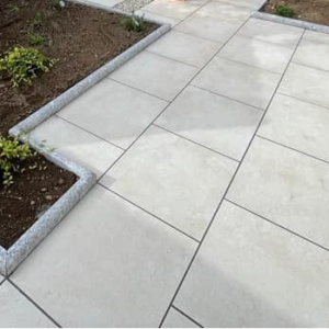 A neatly laid geometric pattern of large square paving stones creating a pathway, bordered by concrete edges, with surrounding beds of young shrubs and plants.