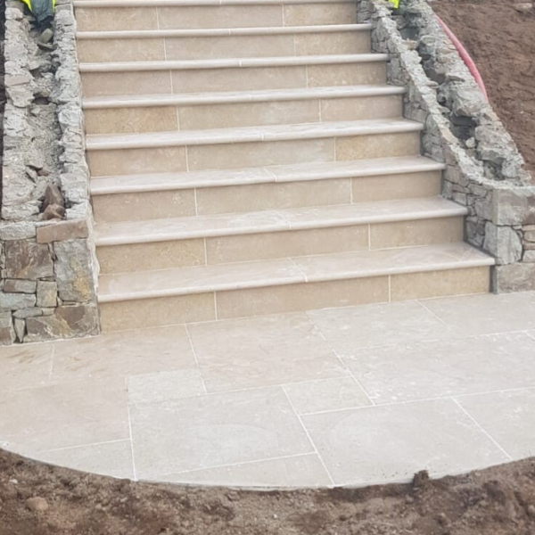 A newly constructed outdoor staircase with stone steps, flanked by stone walls, leading up to an unseen destination. The landing at the bottom is paved with large tiles, and the surrounding ground appears to be freshly worked earth.