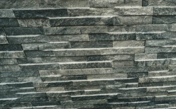 A textured wall made up of stacked grey stone slabs with varying shades and patterns. A sign that reads "NATURA LAVA" is affixed to the wall. The base of the wall is surrounded by small multicolored pebbles.