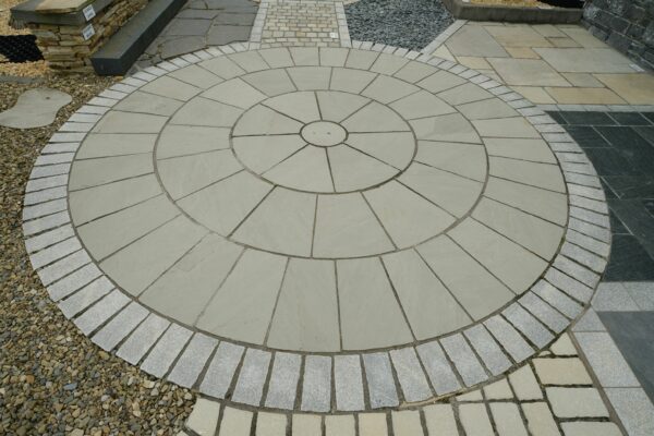 A circular patterned paving design with concentric circles made from cut stone slabs, bordered by a curved row of rectangular pavers and surrounded by gravel and contrasting darker stone pathways.