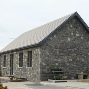 A stone building on a landscaped area with various paving patterns, raised beds, and shrubs, under a cloudy sky.