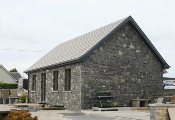 A stone building on a landscaped area with various paving patterns, raised beds, and shrubs, under a cloudy sky.