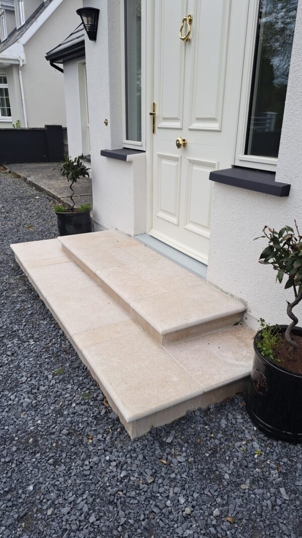 A set of three stone steps leading to a white front door with golden hardware, flanked by a small plant in a black pot, surrounded by gravel landscaping.