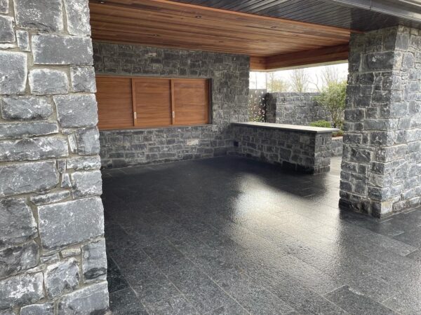 A modern outdoor space with stone columns and walls, featuring wooden panels and a stone counter, under a wooden ceiling. The floor is sleek and shiny, reflecting the light from the entrance.