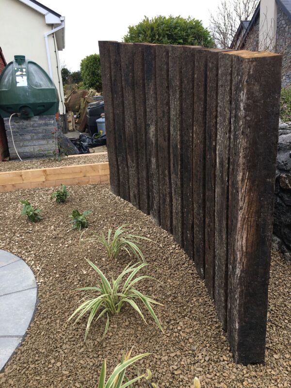 A wooden fence made from dark, rough timber planks is positioned in a garden area featuring pebbled ground and green plants, with various garden supplies and a green water tank visible in the background.