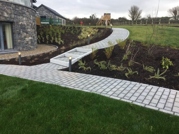 A landscaped pathway made of gray pavers winds through a garden featuring low shrubs and plants, with a stone building in the background. Soft lighting fixtures line the pathway, leading to a grassy area and a play structure in the distance.