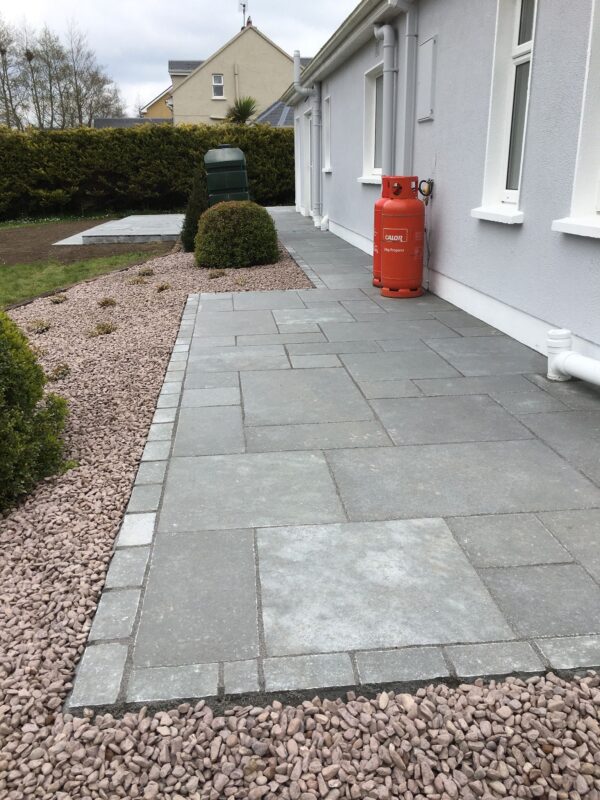 A well-maintained patio area made of gray stone tiles, bordered by small gravel, with two rounded shrubs. An orange gas cylinder is positioned against the wall of a white house, along with a green water storage tank in the background.