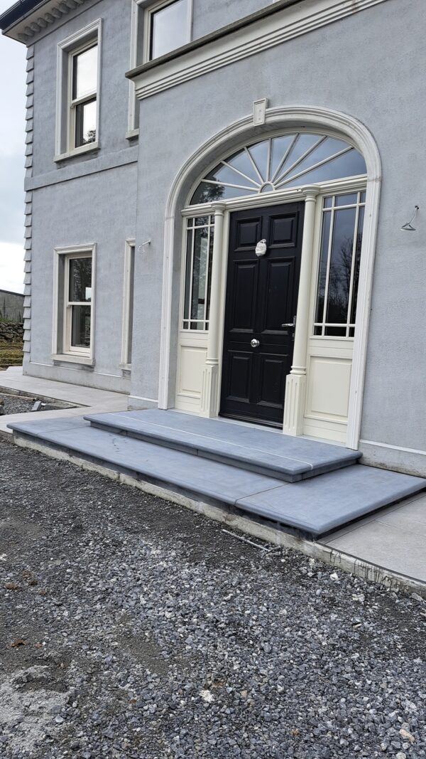 A modern entrance featuring a black front door with decorative molding, framed by large glass panes, and flanked by vertical columns. The entryway includes a set of gray stone steps leading up to the door, with surrounding gravel and concrete work visible.