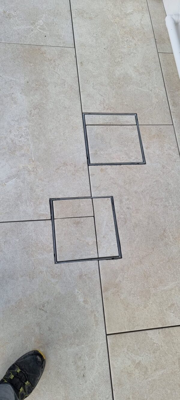 A close-up view of a tiled floor featuring two rectangular black frames set into the tiles, with a shoe partially visible in the lower corner.