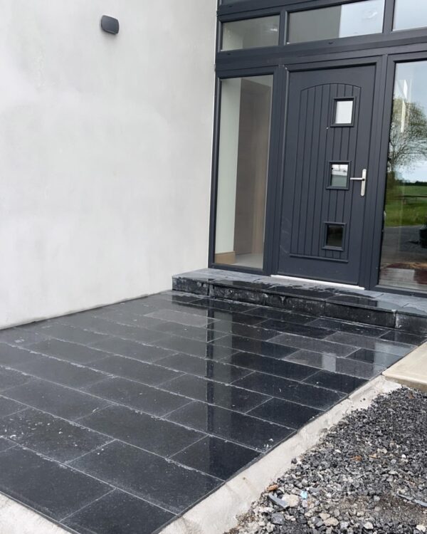 A modern entryway featuring a black tiled porch and a dark wooden door with rectangular windows, framed by a smooth gray wall. There is a small set of steps leading up to the door, and the ground is uneven, with exposed gravel on one side.