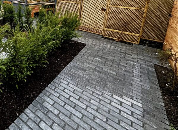 A paved pathway made of gray bricks, bordered by lush green shrubs, with a wooden lattice fence in the background.