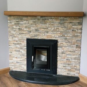 A modern fireplace with a black metal frame and a glass front, set into a textured stone wall, topped with a wooden mantel, against a light-colored wall, with a curved black stone base and wooden floor.