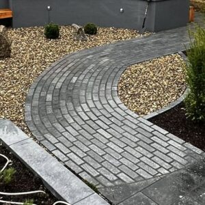 A curved stone pathway made of gray pavers winds through a landscaped garden with small bushes and gravel. Decorative rocks are placed among the gravel, and a layer of dark mulch borders the pathway.