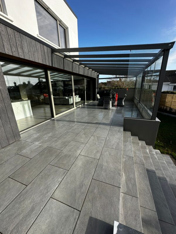 A modern patio with large gray tiles, featuring a glass-covered area with seating and steps leading down to a grassy area, under a clear blue sky.