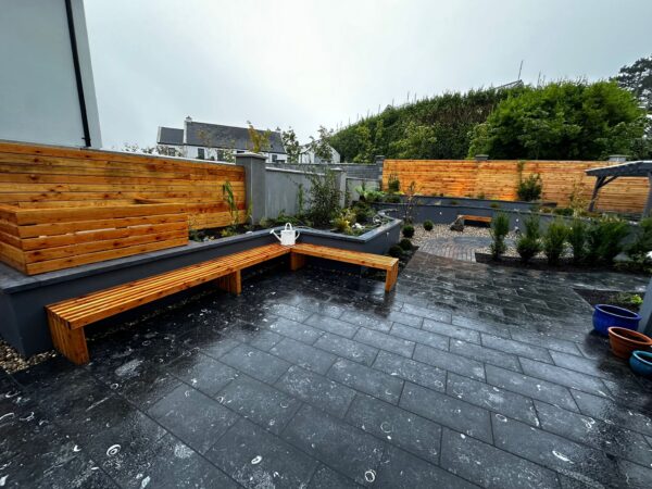 A modern garden featuring wooden benches, stone paving, and landscaped areas with plants. There are wooden panels along the back and a watering can placed on one bench, with light rain causing puddles on the ground.