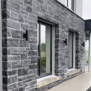 Exterior view of a building featuring a textured gray stone wall with two large windows framed in black, complemented by modern black wall-mounted lights. A concrete walkway leads to the entrance.