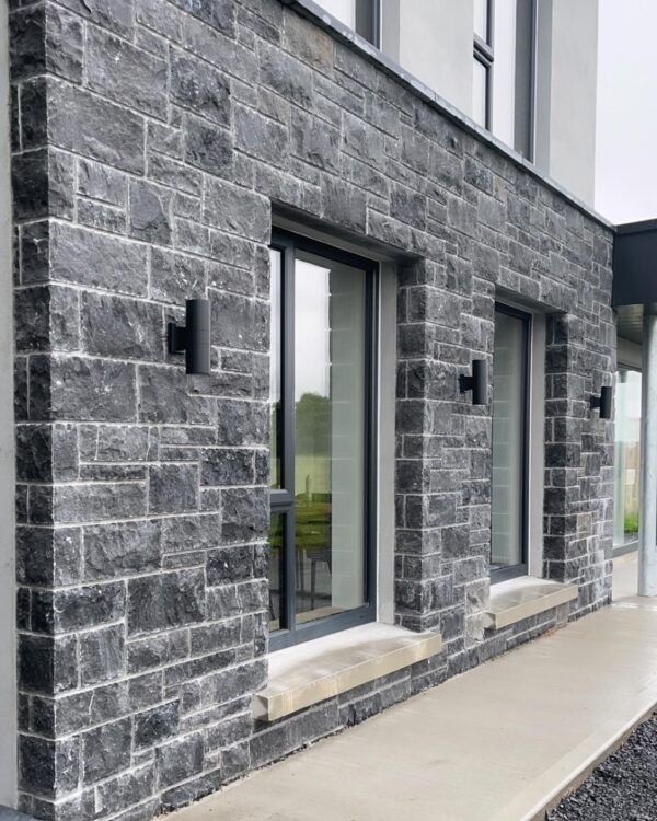 Exterior view of a building featuring a textured gray stone wall with two large windows framed in black, complemented by modern black wall-mounted lights. A concrete walkway leads to the entrance.