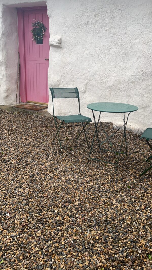 A quaint outdoor seating area features two green metal chairs and a matching round table on a gravel surface, with a white cottage wall and a pink door adorned with a wreath in the background.