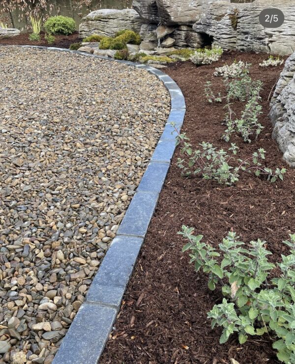 A curved stone border separates a gravel area from a mulched garden bed, featuring small green plants and decorative rocks in the background.